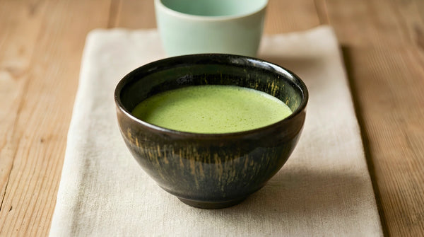 A dark tenmoku-style teabowl of bright green matcha on linen, with a pale celadon cup blurred behind.