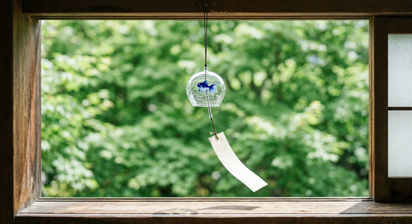 A clear Edo-glass goldfish wind chime with a paper strip hanging in a sunlit open window framed by green foliage.