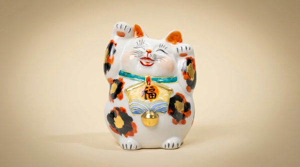 Un maneki neko Kutani de tres colores pintado a mano con la pata levantada, sobre un fondo cálido y suave.