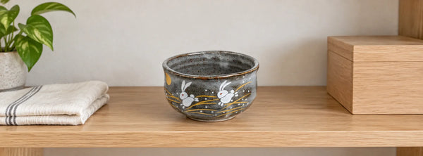 Grey Kutani rabbit matcha bowl on a wooden shelf beside a folded cloth and a plain storage box.