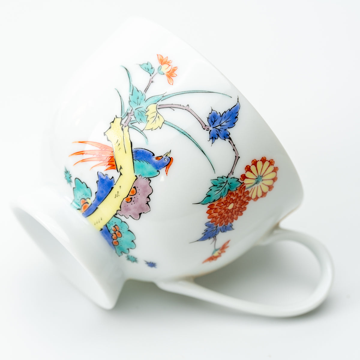 Chrysanthemum and bird cup resting on its side, showing colorful flowers, a yellow branch and a white handle.