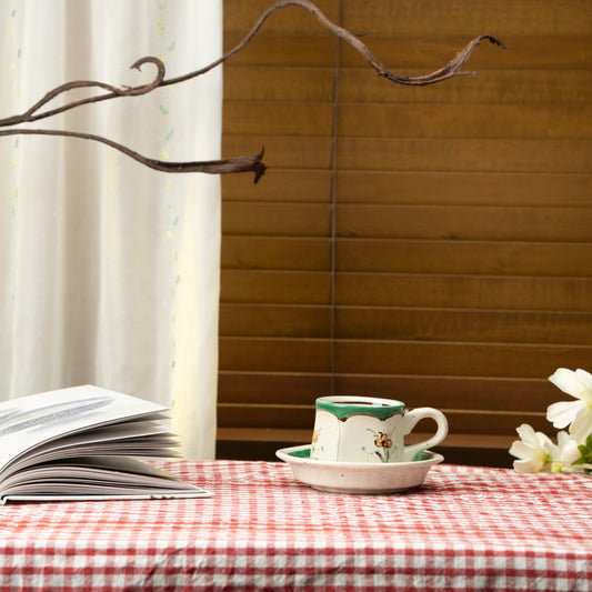 Green floral demitasse cup and saucer on a red checked tablecloth beside a book.