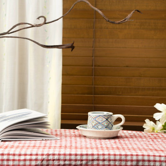 Diamond-pattern cup and saucer on a red checked tablecloth beside a book and flowers.