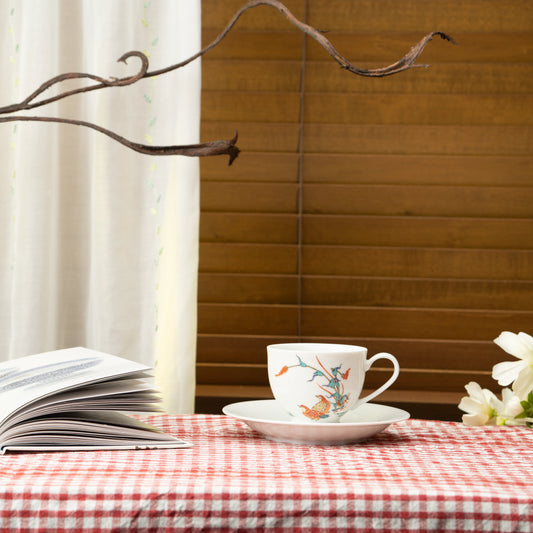 Porcelain cup and saucer with quail and red berries on a red checked tablecloth beside a book.