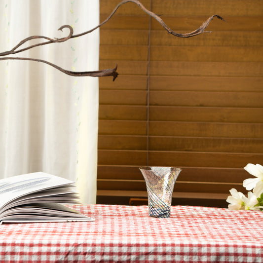 Blue and gold-colored Yachiyo glass sake cup on a red checked tablecloth beside a book.