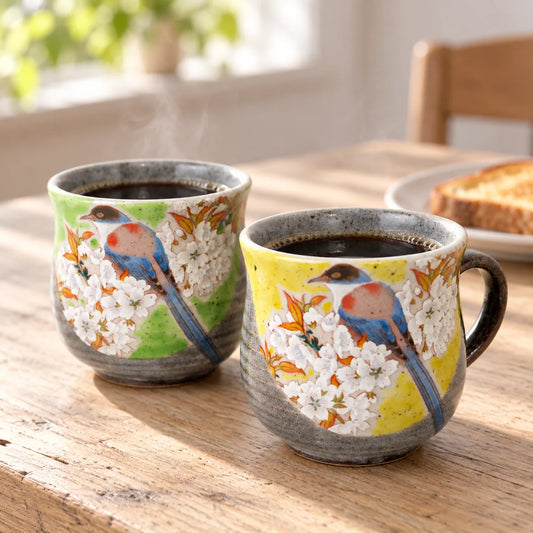 Two Kutani bird and cherry blossom mugs, one green and one yellow, filled with coffee on a sunlit breakfast table