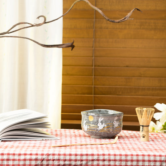 Leaping rabbits matcha bowl on a red checked tablecloth beside a bamboo whisk.
