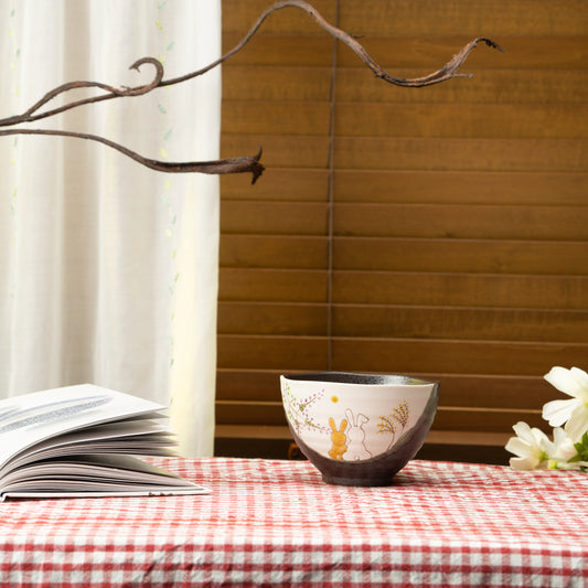 Rabbit bowl on a red checked tablecloth beside a book and flowers.