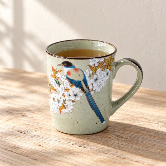 Sage-green Kutani mug with a blue bird and white cherry blossoms, holding tea on a sunlit wooden table.