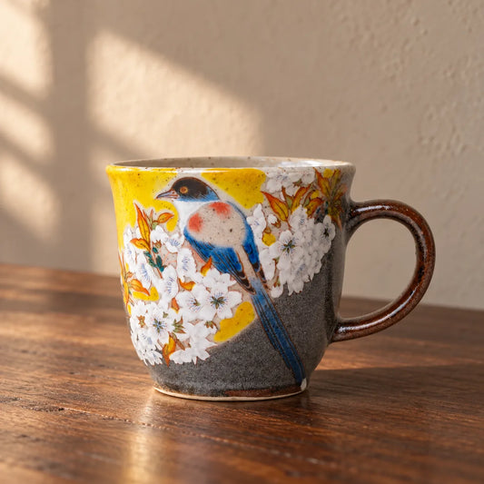 Yellow and charcoal Kutani mug painted with a blue bird and white cherry blossoms on a walnut table.