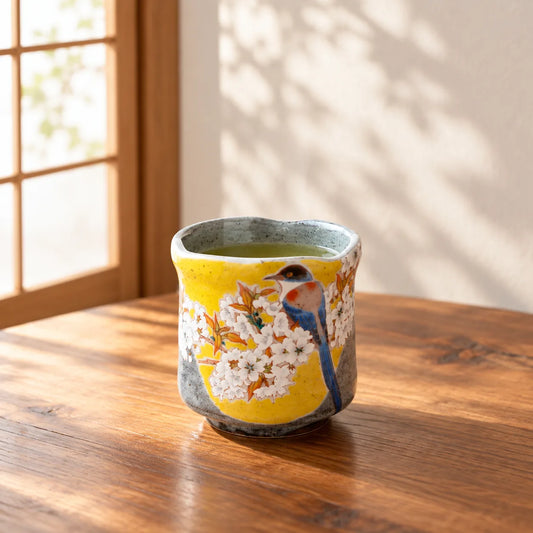 Kutani Yoshidaya yunomi with blue bird and white cherry blossoms holding green tea by a window