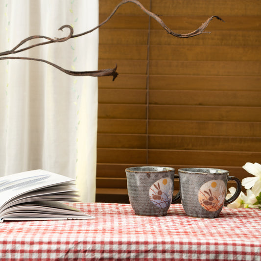 Rabbit mug pair on a red checked tablecloth beside a book and white flowers.