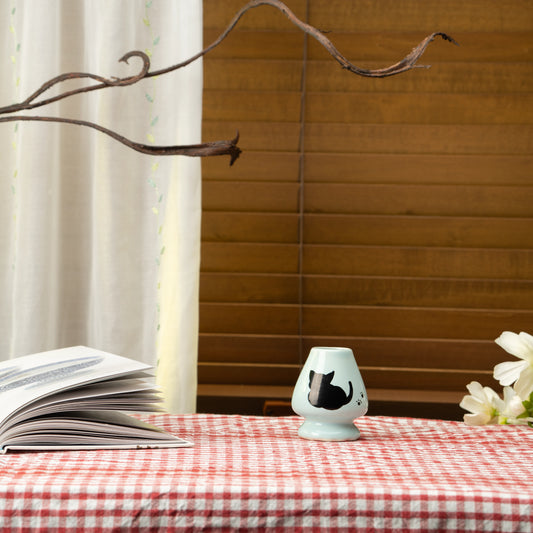 Pale blue cat whisk holder on a red checked tablecloth beside a book and flowers.