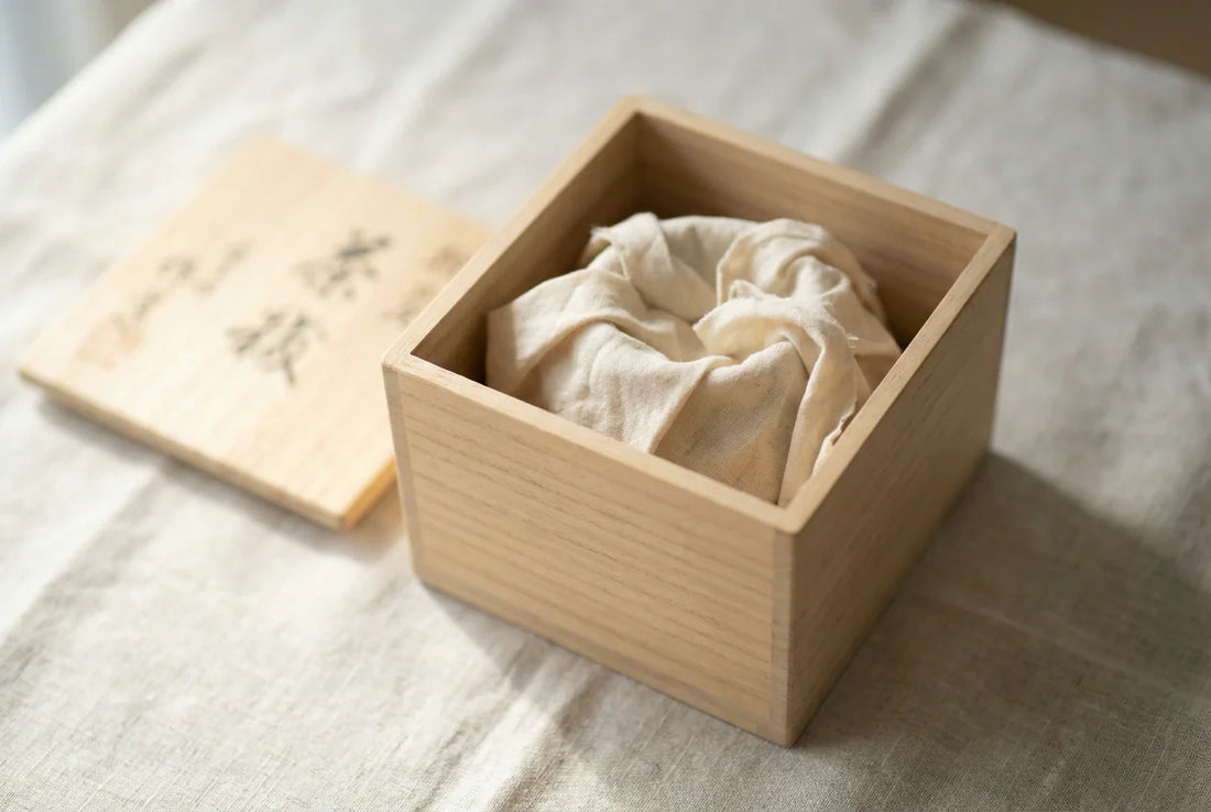 An open paulownia tomobako box with a brushed-calligraphy lid and a cotton-wrapped ceramic piece inside on linen