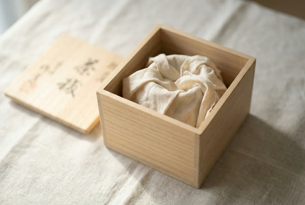 An open paulownia tomobako box with a brushed-calligraphy lid and a cotton-wrapped ceramic piece inside on linen