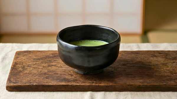 A black kuro-raku-style hand-molded Japanese tea bowl with whisked matcha on linen by a shoji screen.