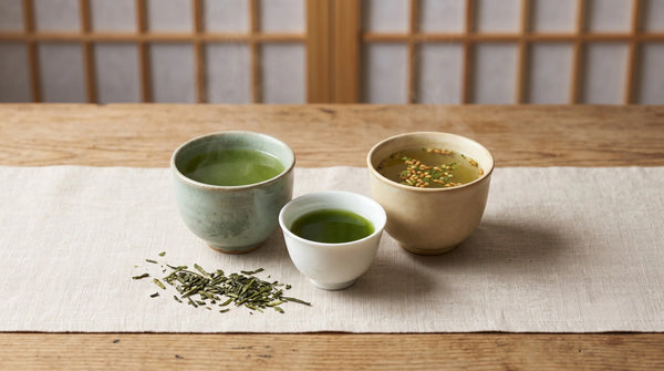 Three small Japanese cups of Shizuoka green tea — cloudy sencha, thick jade gyokuro, and golden genmaicha — with loose leaf on linen.