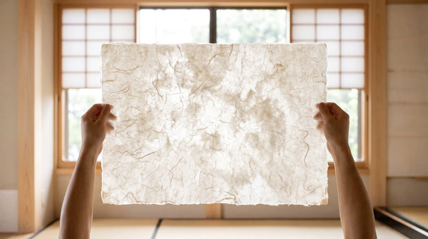 Hands holding a translucent sheet of washi up to window light, long fibers visible in the paper