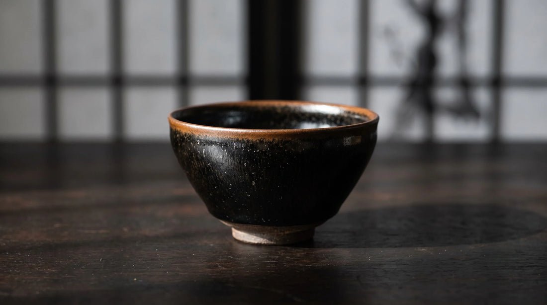A glossy black tenmoku tea bowl with a persimmon-brown rim and faint oil-spot flecks on dark wood