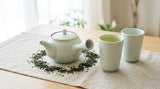 A pale-celadon side-handle kyusu teapot with two yunomi cups of green sencha on linen by a window.