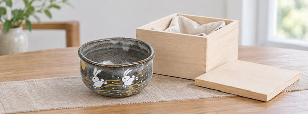 Grey Kutani rabbit matcha bowl beside an open wooden box with soft cloth on a dining table.