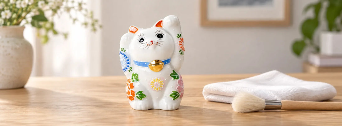 White floral Kutani lucky cat beside a clean folded cloth and a soft dusting brush on a wooden table.