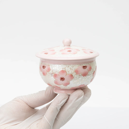 Pink cherry blossom teacup with its lid on, held in a gloved hand to show the cup’s size and rounded shape.