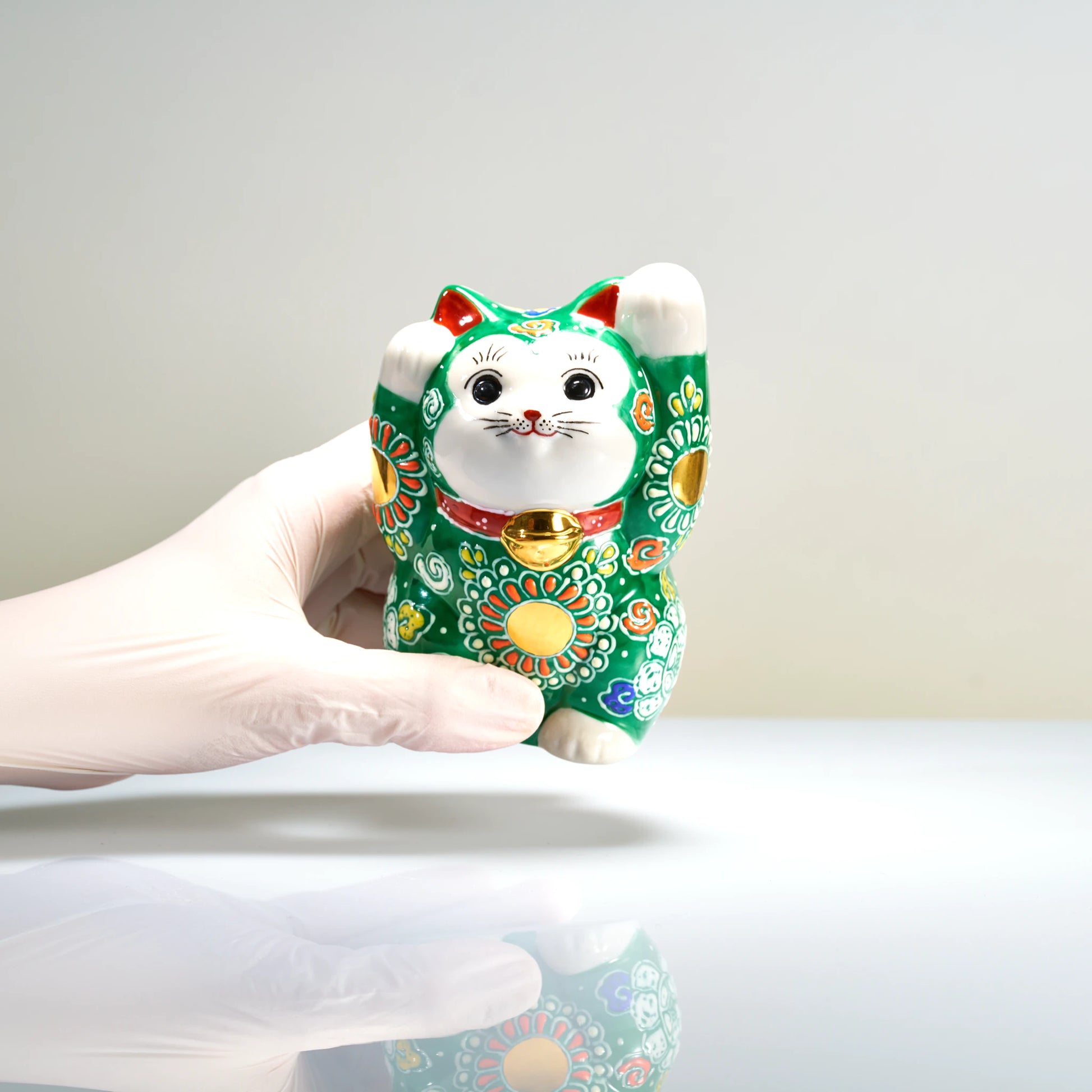A gloved hand holding the 12 cm tall green Kutani beckoning cat to demonstrate its size and scale.