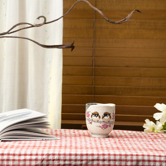 Sparrow yunomi on a red checked tablecloth beside an open book and white flowers.
