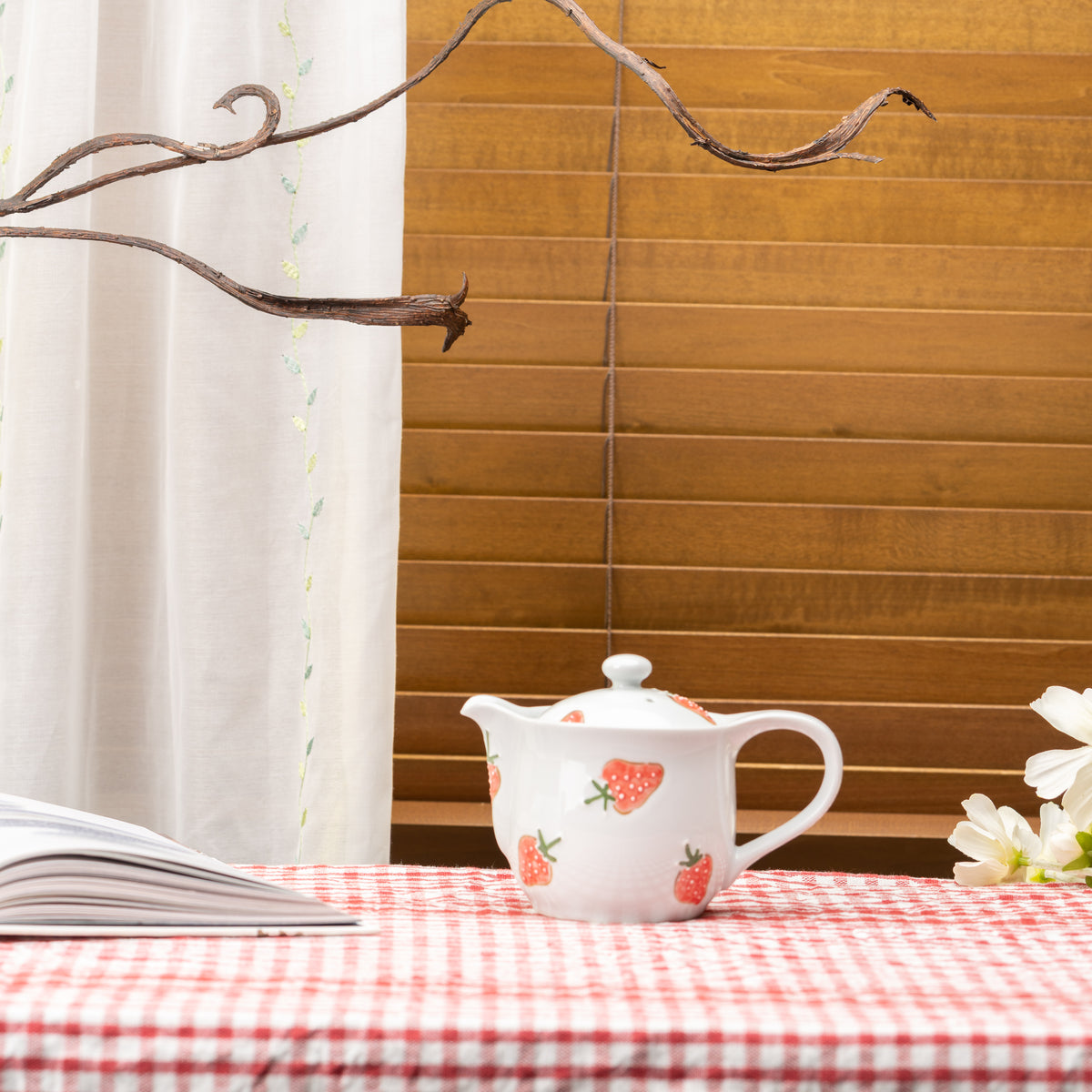 Strawberry teapot on a red gingham tablecloth beside an open book and white flowers.