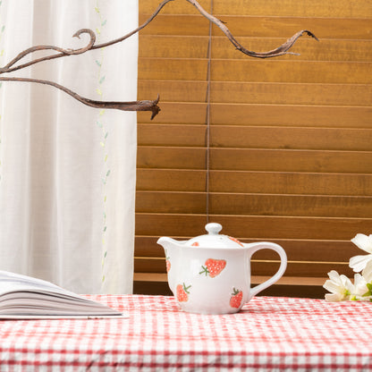 Strawberry teapot on a red gingham tablecloth beside an open book and white flowers.