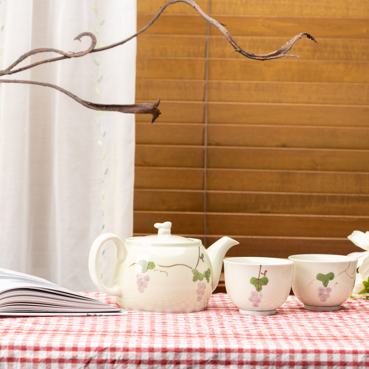 Grape-patterned Arita teapot and two cups displayed on a red gingham tablecloth beside an open book.