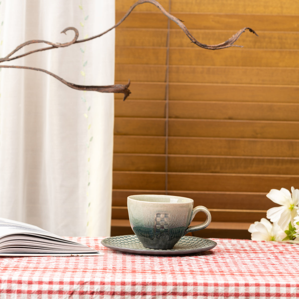 Hechimon Mosaic cup and saucer on a red gingham tablecloth beside an open book and white flowers.