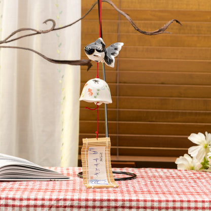 Black goldfish wind chime hanging from a branch, with a white painted bell and bamboo-backed tag below.