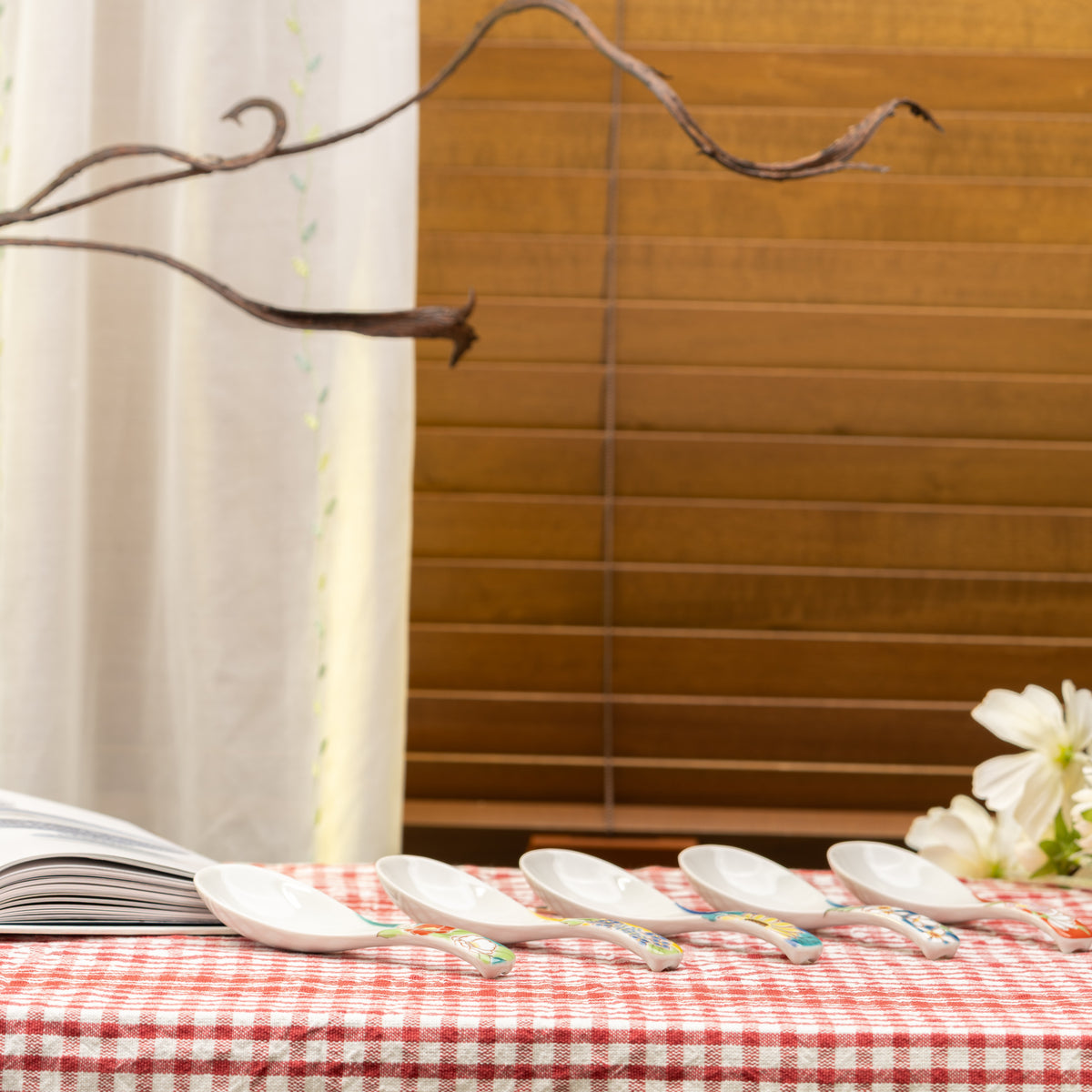 Five floral Kutani soup spoons arranged on a red gingham table beside an open book and white flowers.