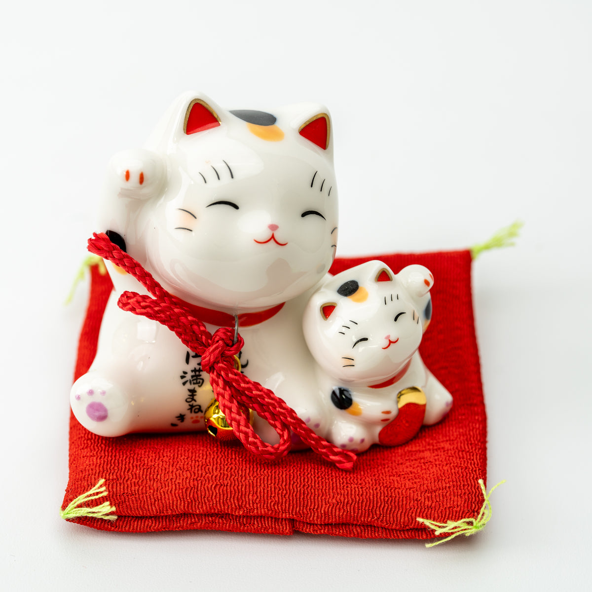 Front view of a calico parent-and-kitten lucky cat with red cords, seated on a red cushion.