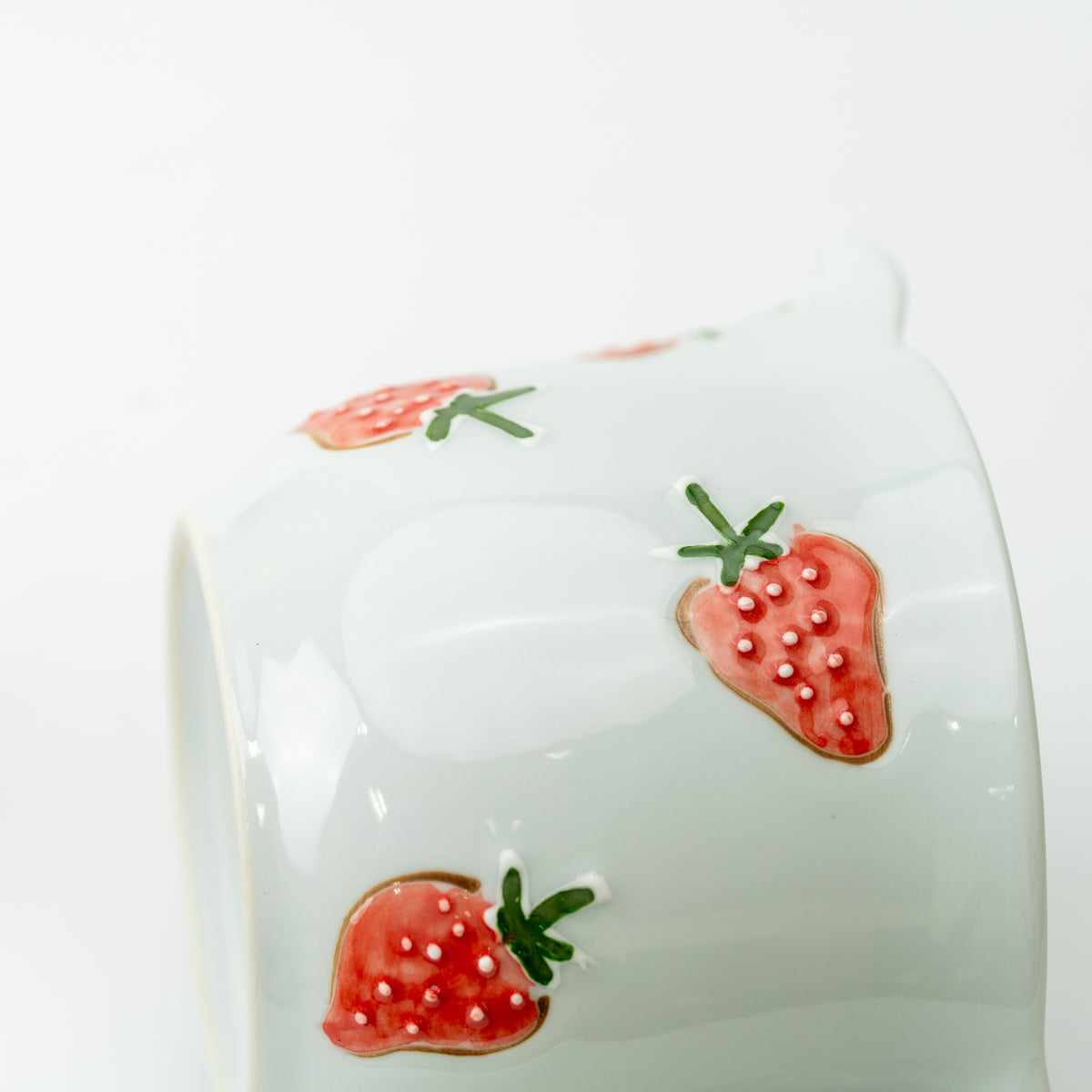 Close-up of raised red strawberries and green leaves on the side of the white teapot.