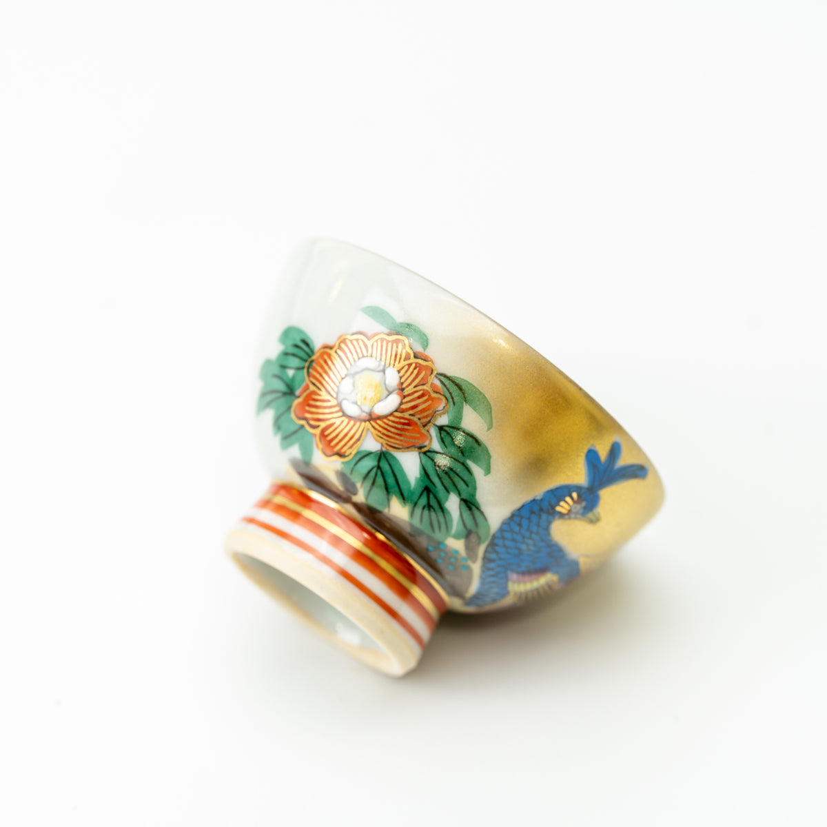 Kutani sake cup tilted on its side, showing a blue peacock, orange peony and striped foot.