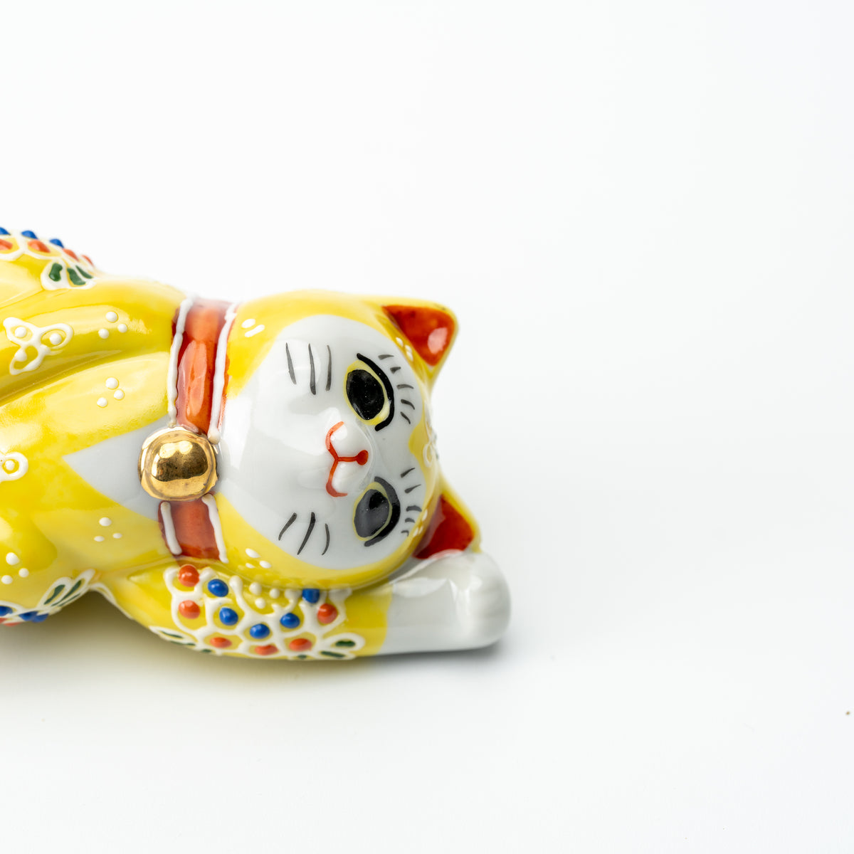 Close-up of the yellow Mori lucky cat's face, red collar and gold-colored bell while lying on its side.