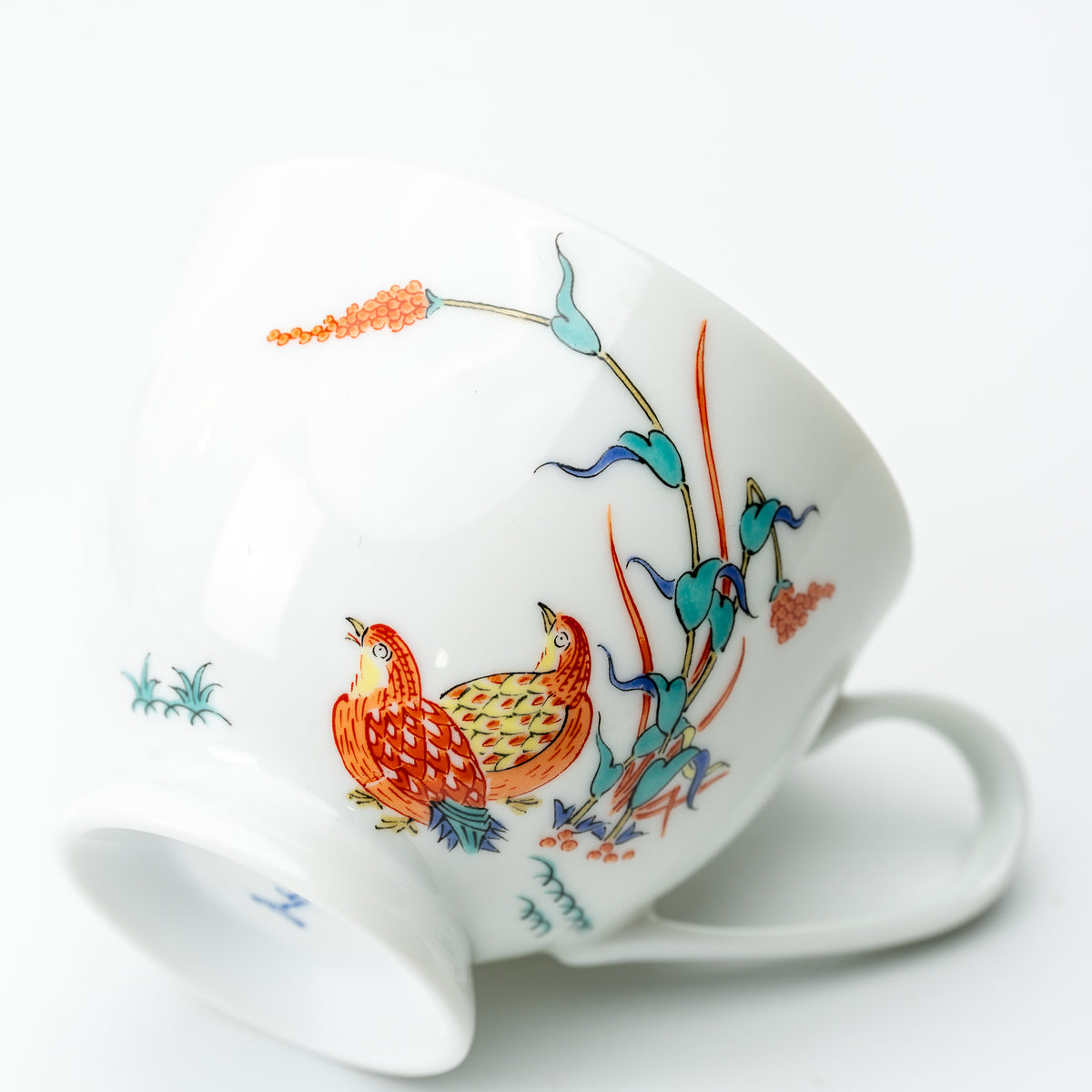 Kakiemon-style cup resting on its side, showing two quail beside colorful grasses and a curved white handle.