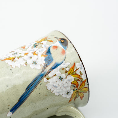 Close-up of a long-tailed blue bird among white cherry blossoms on the pale green Kutani mug.