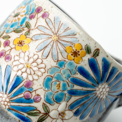 Close-up of the Hana mug's blue flowers with yellow blossoms and purple buds on a pale speckled background.