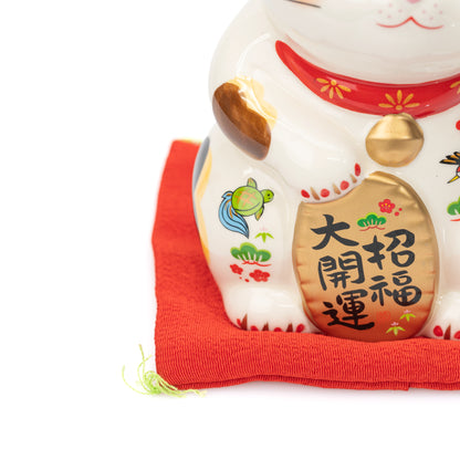 Close-up of a Yakushigama lucky cat's gold-colored plaque, bell and painted decoration above its red cushion.