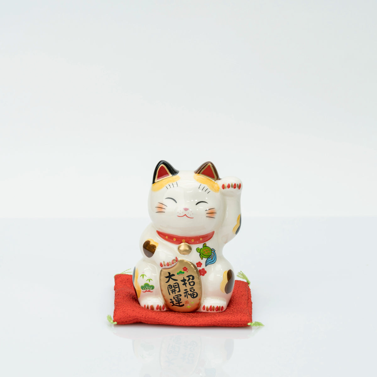 Stationary calico lucky cat seen from the front on its red cushion