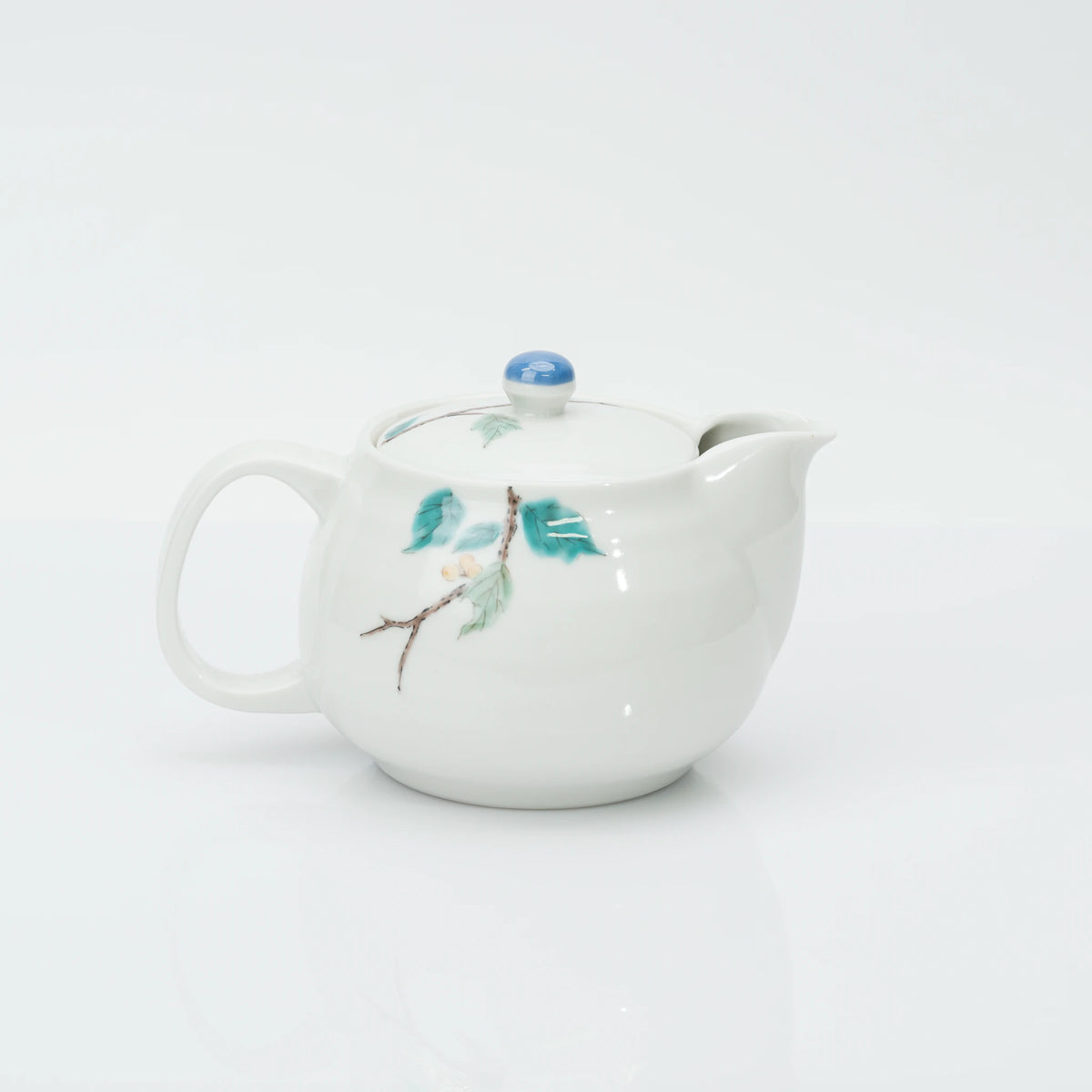 Reverse side of the porcelain teapot, a slim branch with green leaves and a small white blossom, spout to the right.