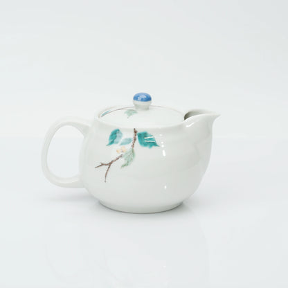 Reverse side of the porcelain teapot, a slim branch with green leaves and a small white blossom, spout to the right.
