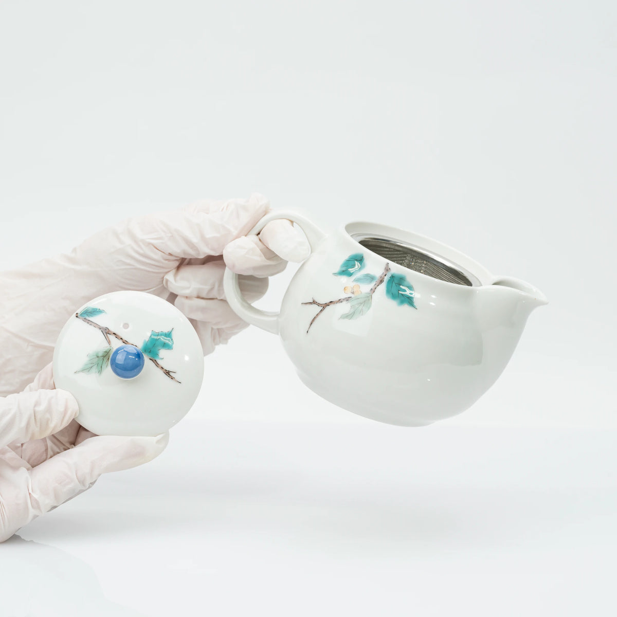 Gloved hands lifting the lid off the teapot, the stainless steel strainer seated inside the wide opening.