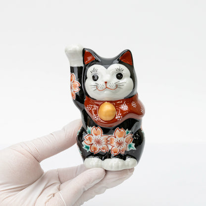 Black Kutani lucky cat held in one hand, showing its raised paw, cherry blossoms, and red bib