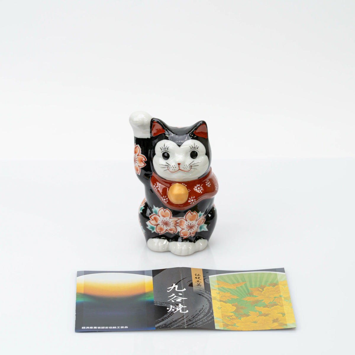 Front view of the black Kutani lucky cat beside a Kutani ware leaflet