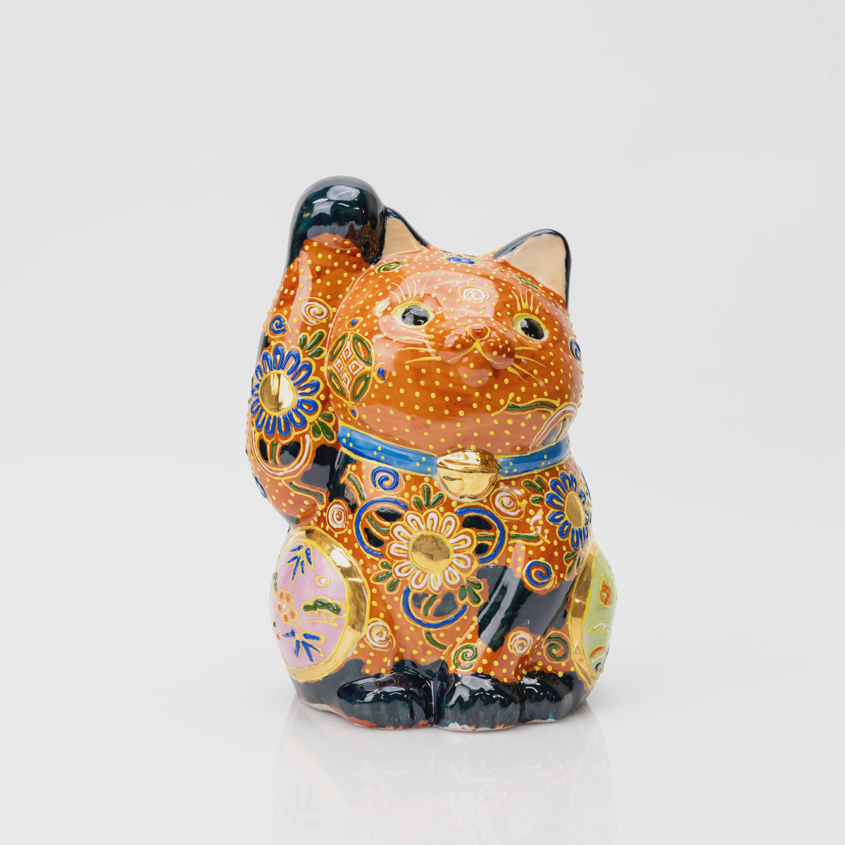 Front view of a Kutani lucky cat with a raised right paw, copper-orange and black coloring, floral patterns, yellow dots and a blue collar.